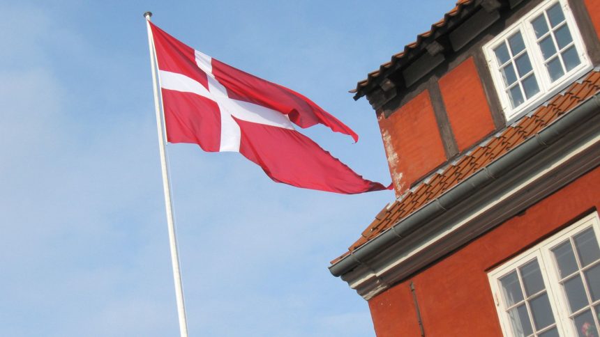Flaga Danii na maszcie na tle nieba i brązowego budynku.