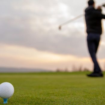 Jaka jest historia golfa? Na zdjęciu piłka do golfa, a w tle mężczyzna robiący zamach kijem do golfa.