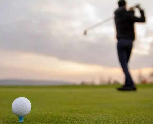 Jaka jest historia golfa? Na zdjęciu piłka do golfa, a w tle mężczyzna robiący zamach kijem do golfa.